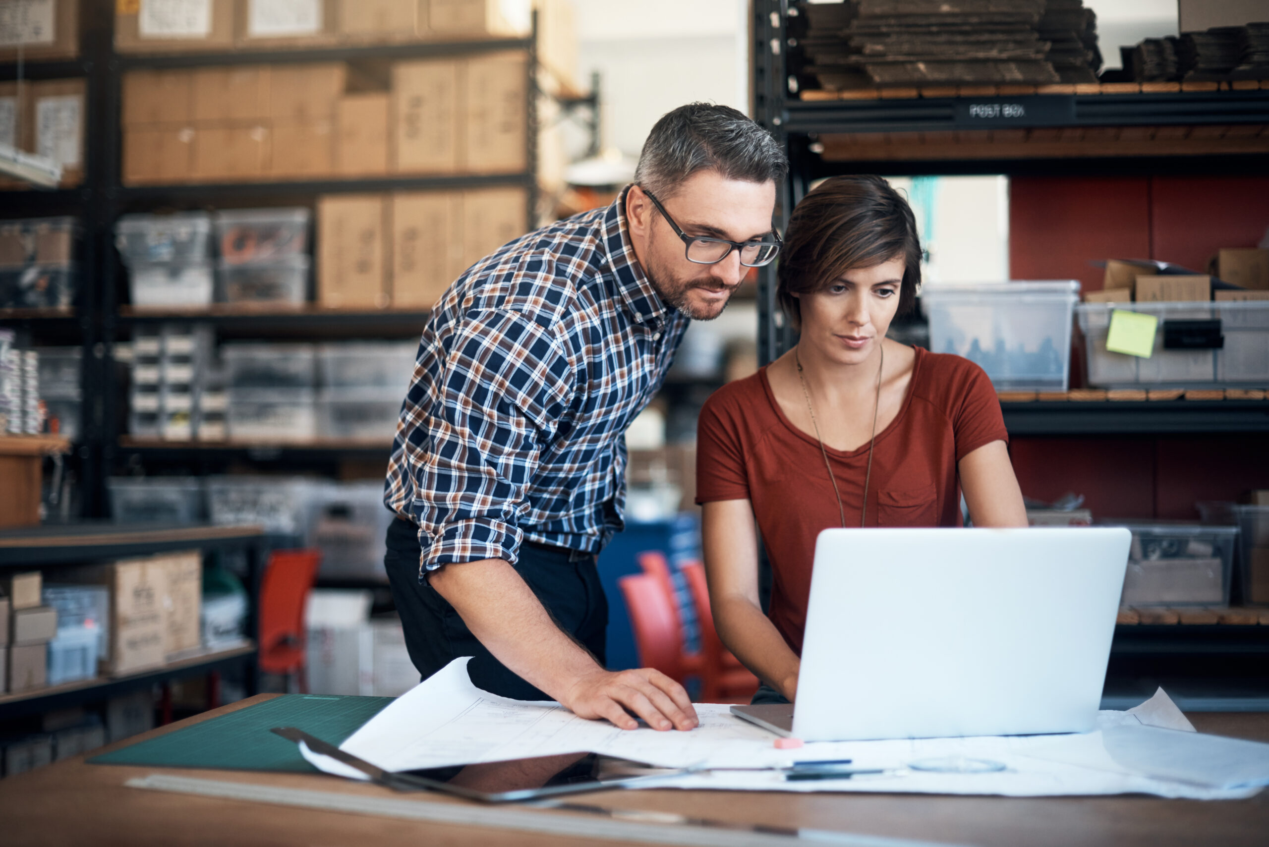 A man and woman working on a laptop at a table in a small business storeroom setting.